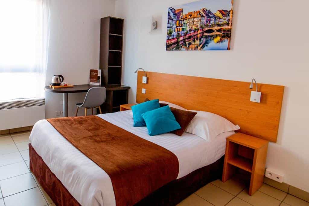 Hotel room with bed, desk, and artwork depicting colorful Strasbourg buildings over water.