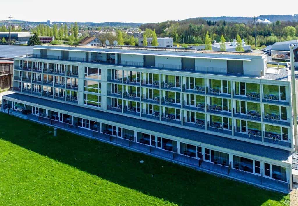 Modern hotel building with many balconies overlooking a bright green lawn and distant trees.