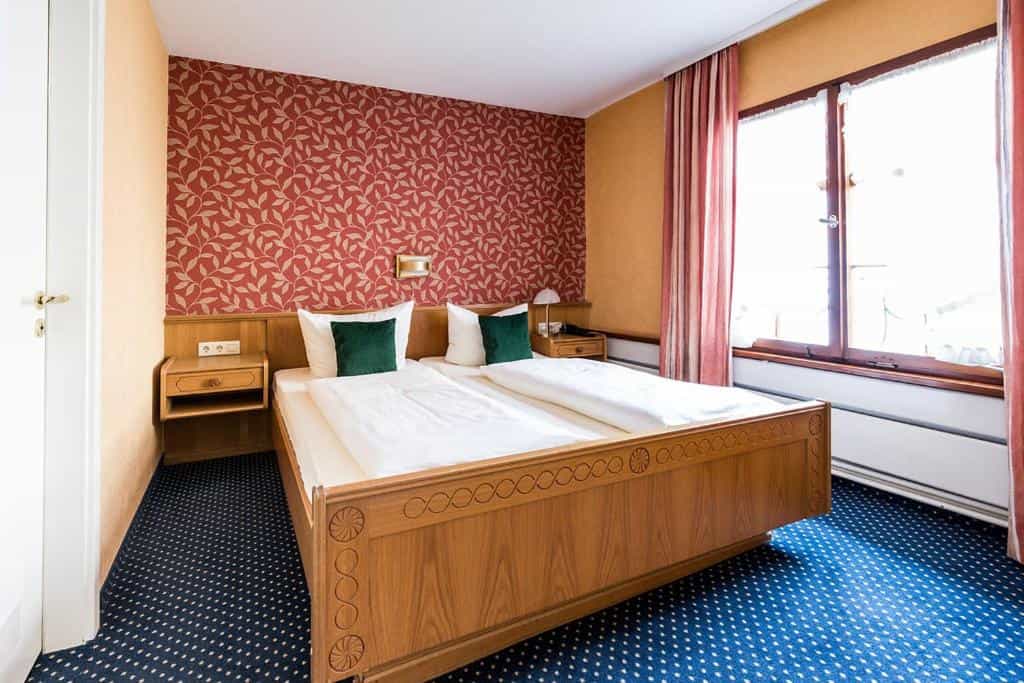 Twin beds with white linens and green pillows in a room with red patterned wallpaper and blue carpet.