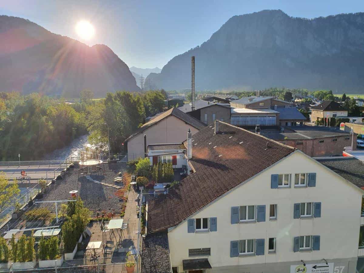 BnB Haus Weibel building with rooftop terrace, river, and sunlit mountains in Buchs.