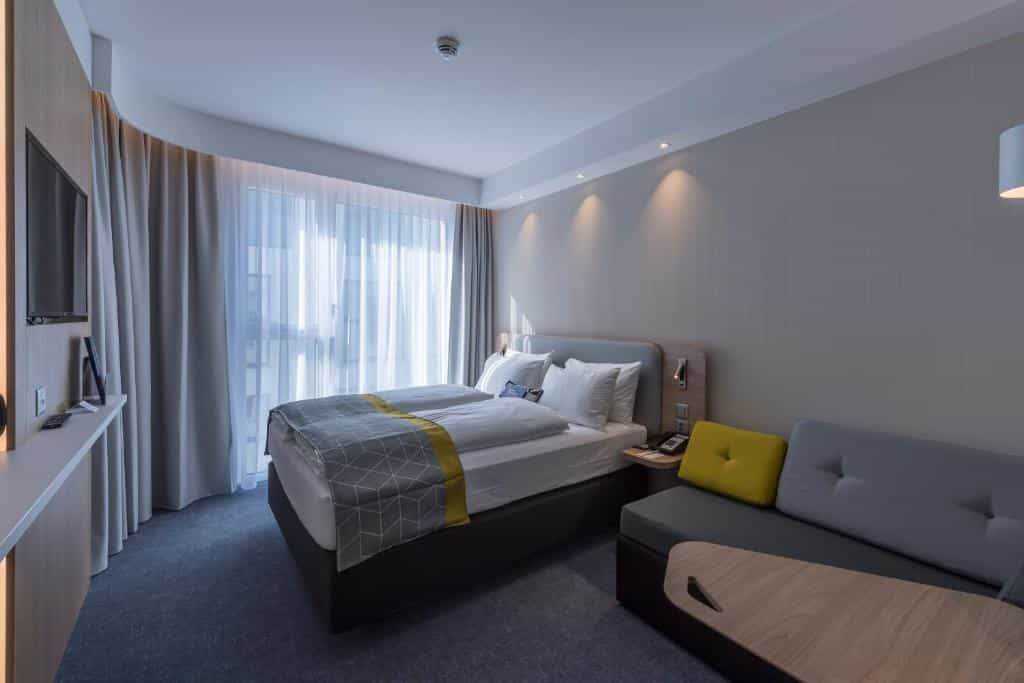 Modern hotel room with twin beds, gray sofa, and sheer window curtains in Lucerne Holiday Inn Express.