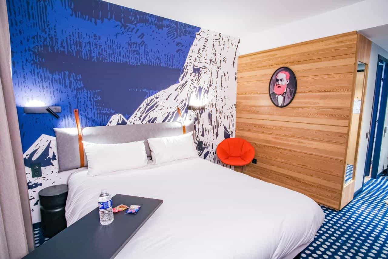 Hotel room with white bedding, blue mountain mural, and wood paneling in Saint-Helene-sur-Isere.