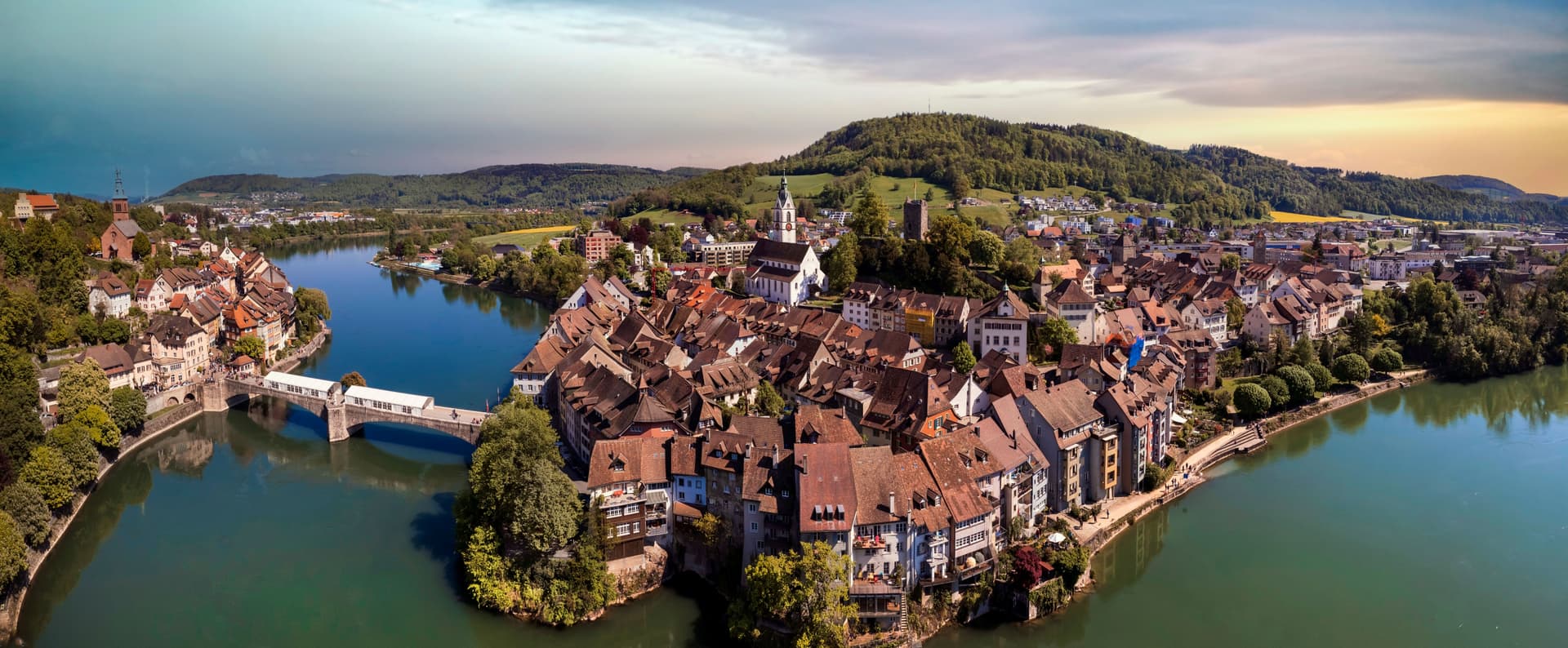Aerial view of Laufenburg town with terracotta roofs, river, and wooded hills under a dramatic sky.