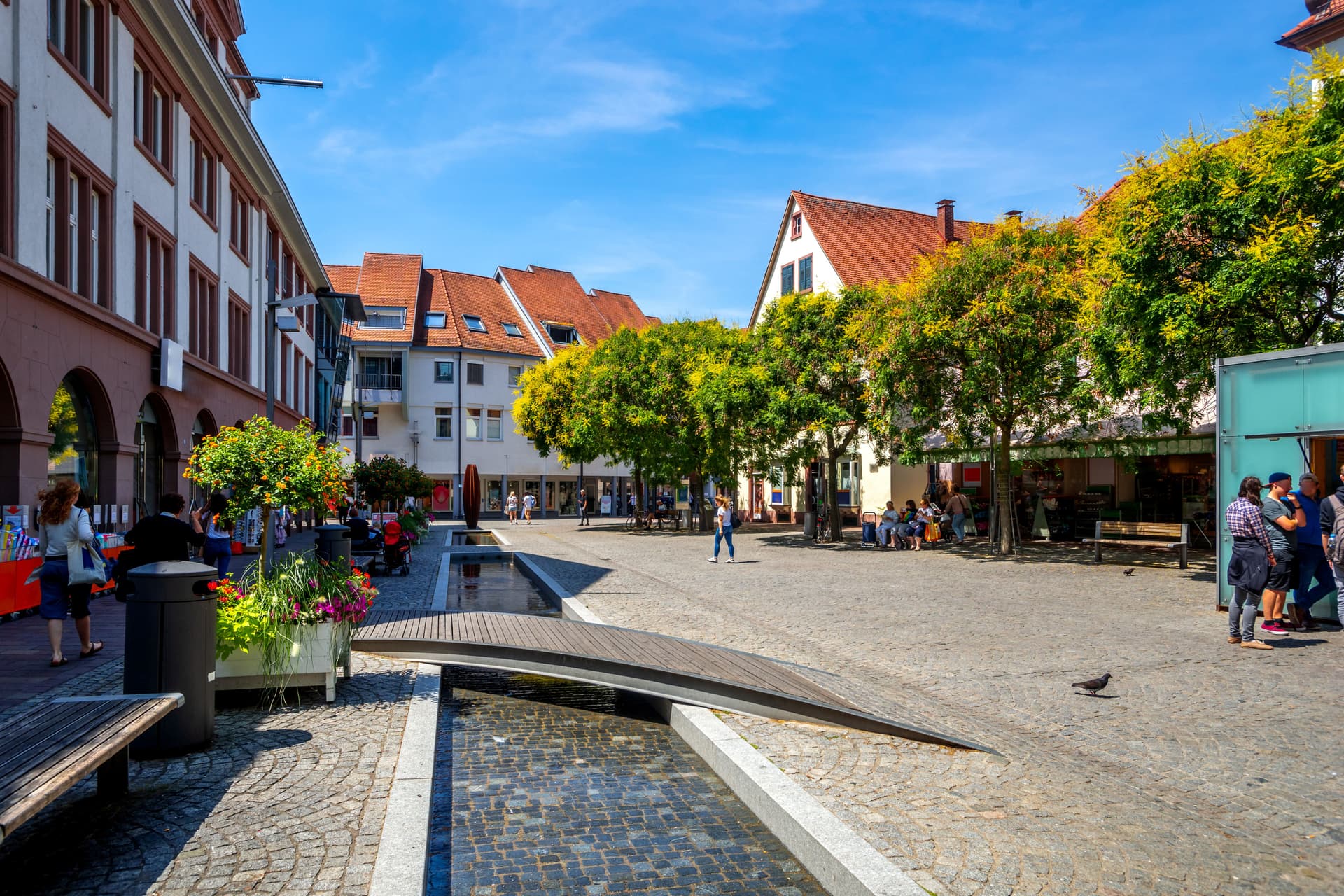 Cobblestone square with water feature, trees, and historic buildings under blue sky in Lahr.