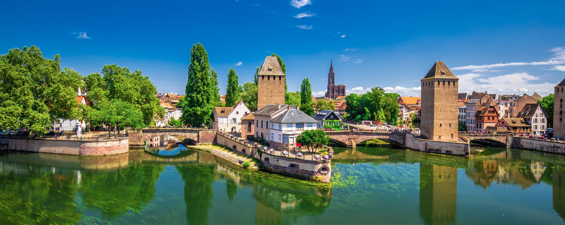 Strasbourg waterfront with historic towers, stone bridges, and green river reflections under blue sky.