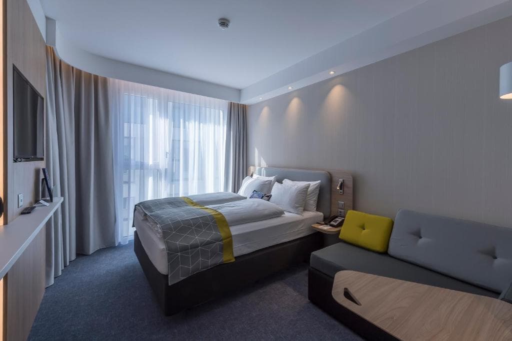 Modern hotel room with twin beds, gray sofa, and sheer window curtains in Lucerne Holiday Inn Express.