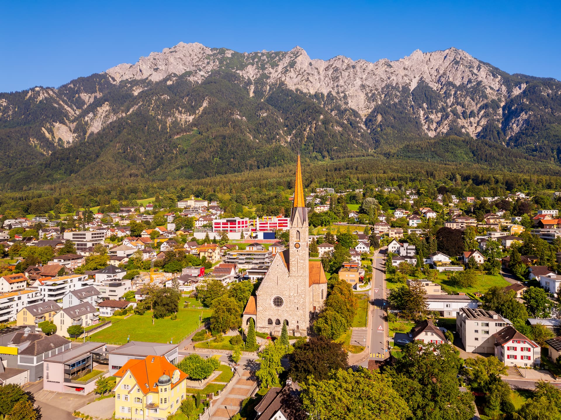 Aerial view of stone church in Schaan, Liechtenstein, set against steep, forested mountains.