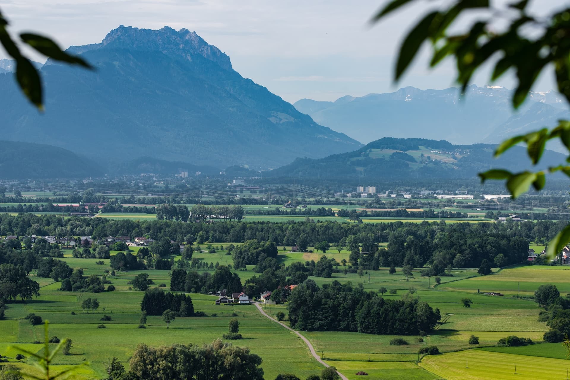 View north from Kummenberg near Koblach over green fields toward hazy blue mountains.