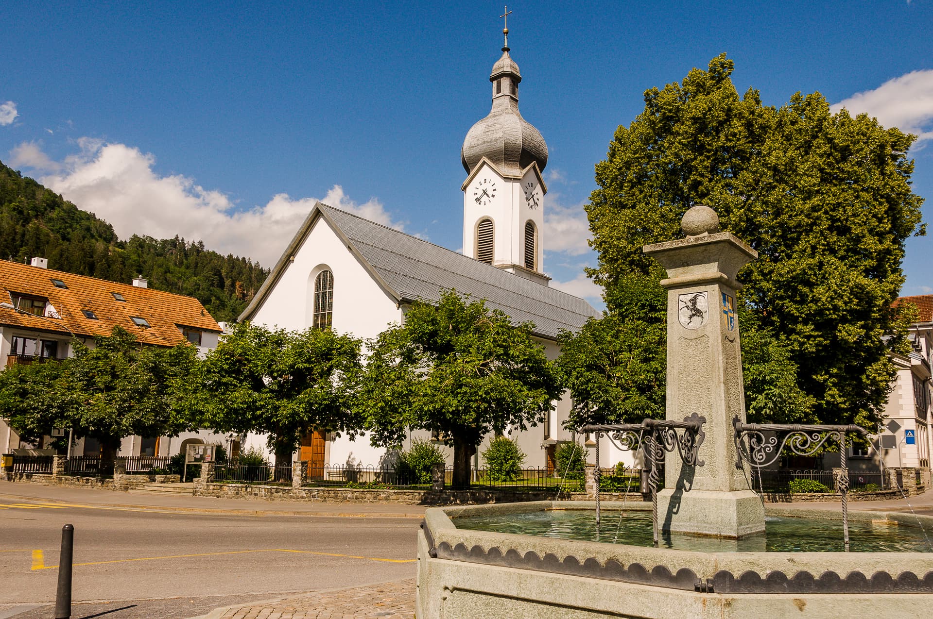 Stone fountain with flowing water in front of white church with clock tower, sunny day.