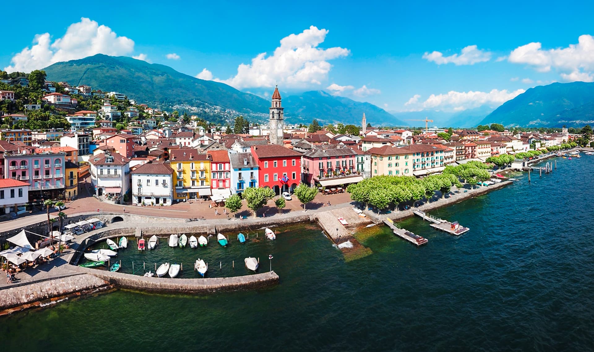 Colorful town on Lake Maggiore with boats docked, backed by steep green mountains.