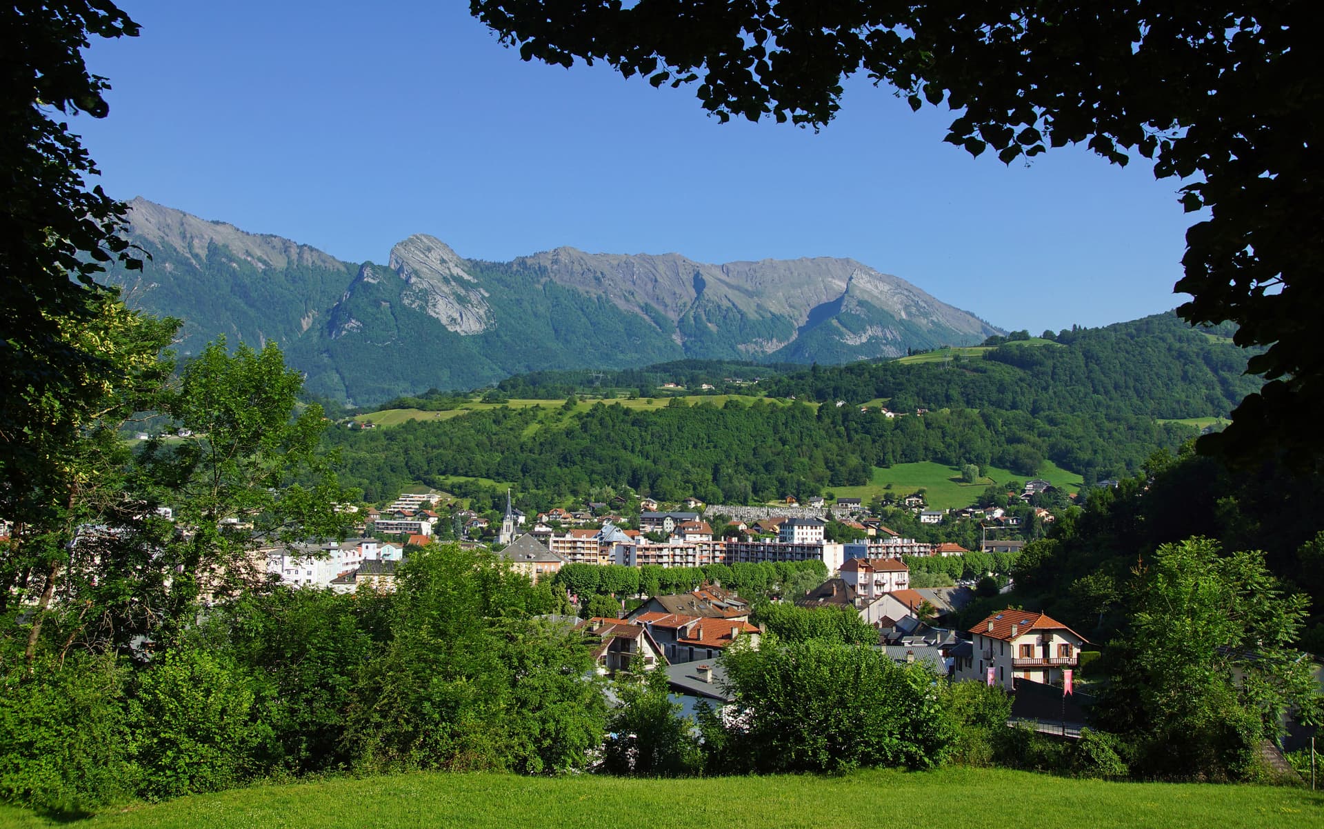 Alpine town nestled in green valley below rugged mountains, viewed through foliage.