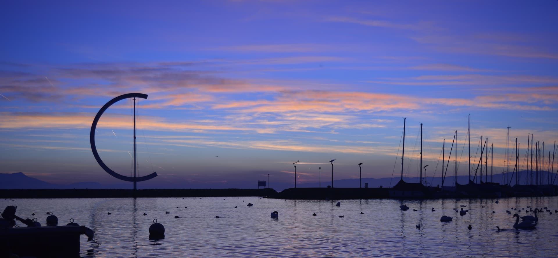 Sunset over Ouchy harbor with swans and sailboat masts on Lake Geneva.