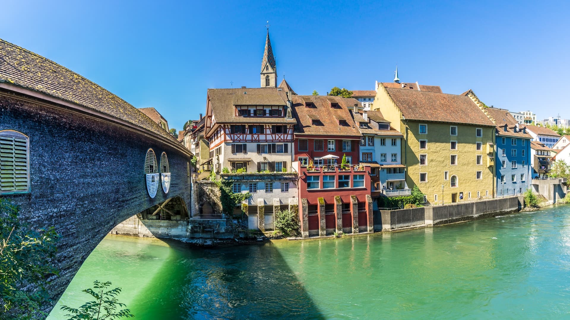 Wooden covered bridge over the green Limmat River next to colorful buildings in Baden, Switzerland.