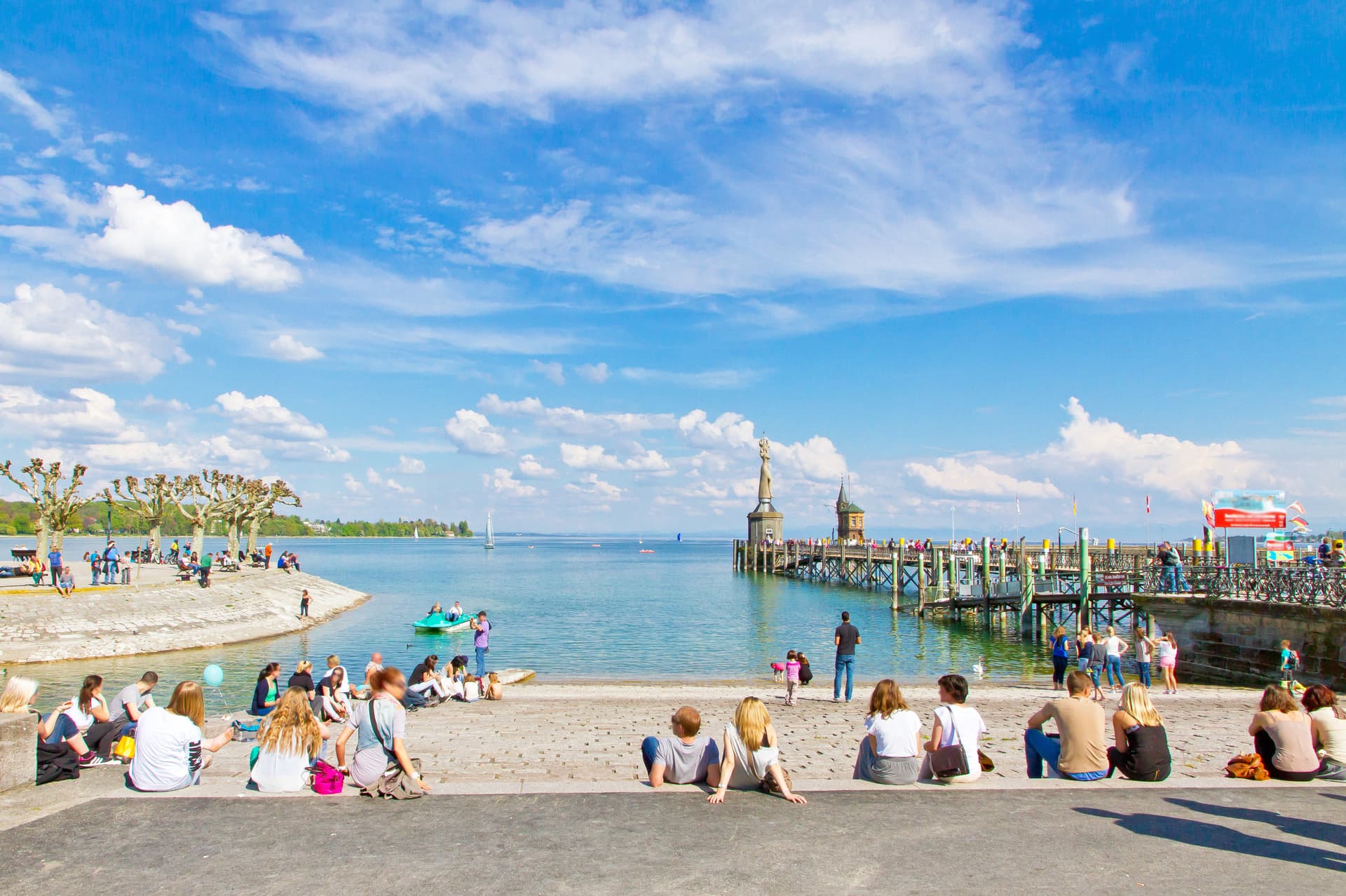 People relaxing by the water near the Lindau harbor statue under a blue sky.