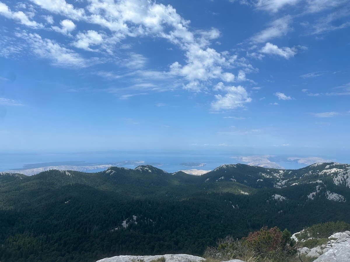 Mountain view overlooking forested slopes, islands, and the sea under a blue sky with clouds.