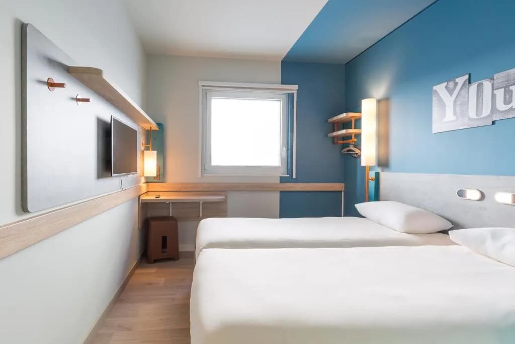 Twin beds in a modern hotel room with blue accent walls at Ibis Geneve Palexpo.