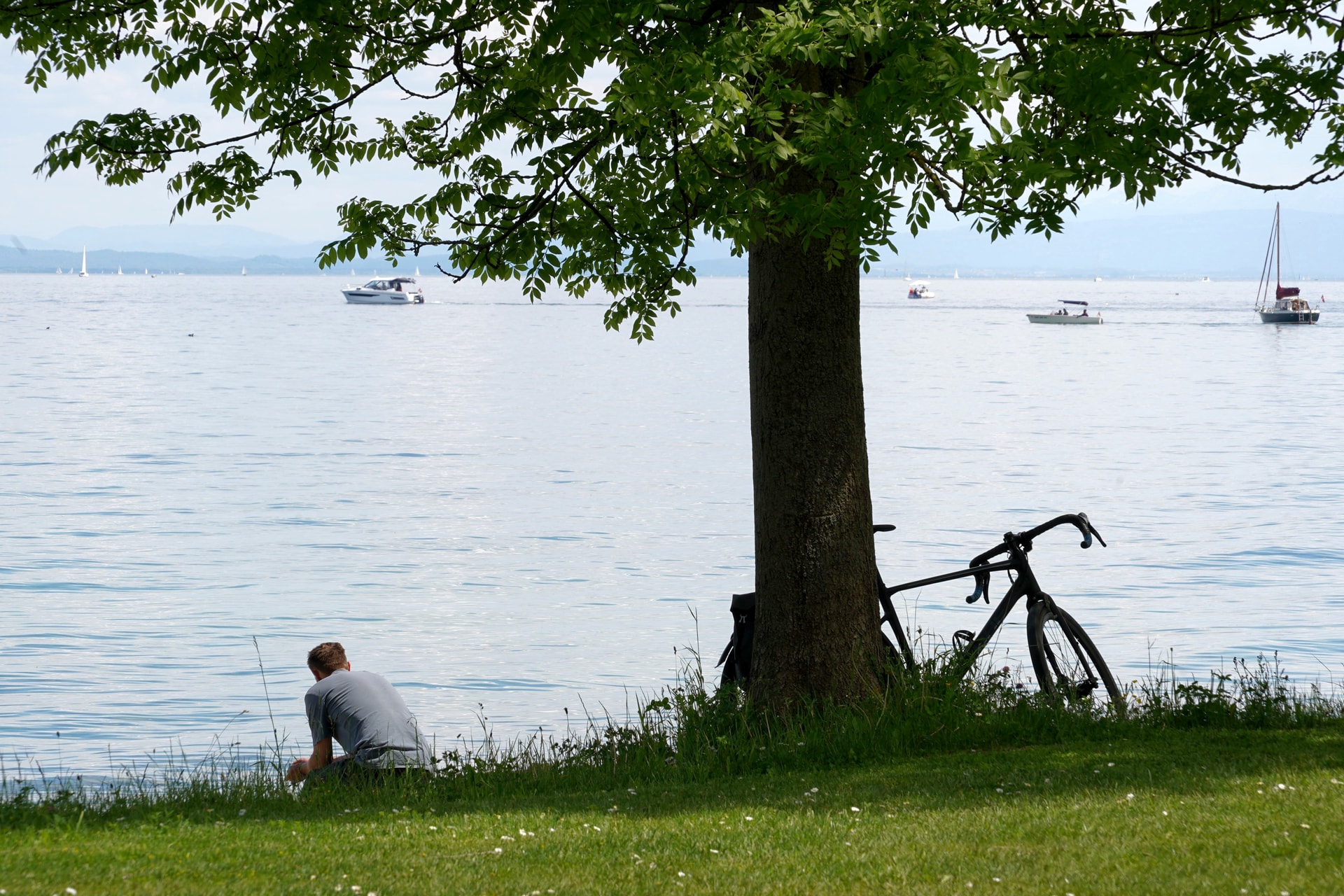 Man sitting by Lake Constance with bicycle, boats on water, and mountains in background.