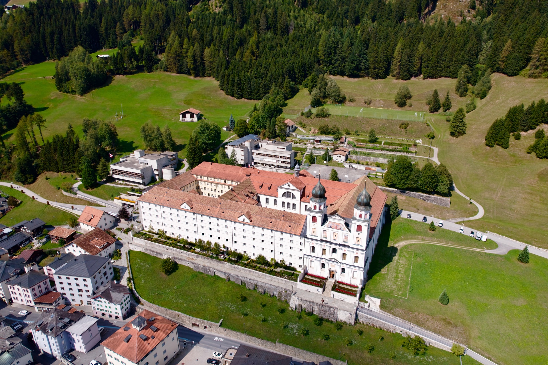 Aerial view of Cloister Disentis with red roofs, white walls, and surrounding green alpine landscape.