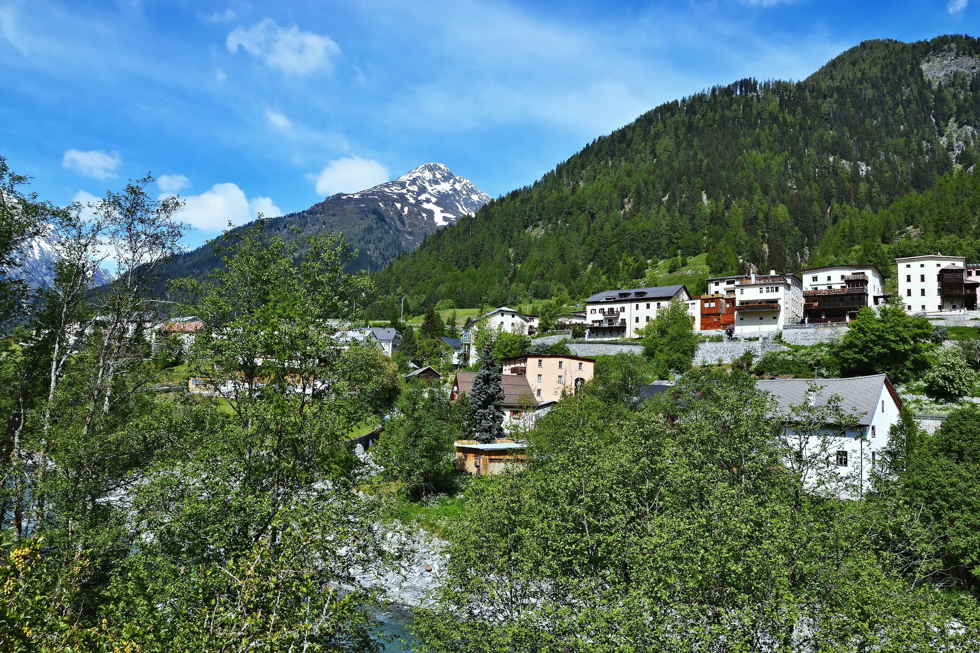 Alpine village of Lavin nestled among green trees with a snow-capped mountain under a blue sky.