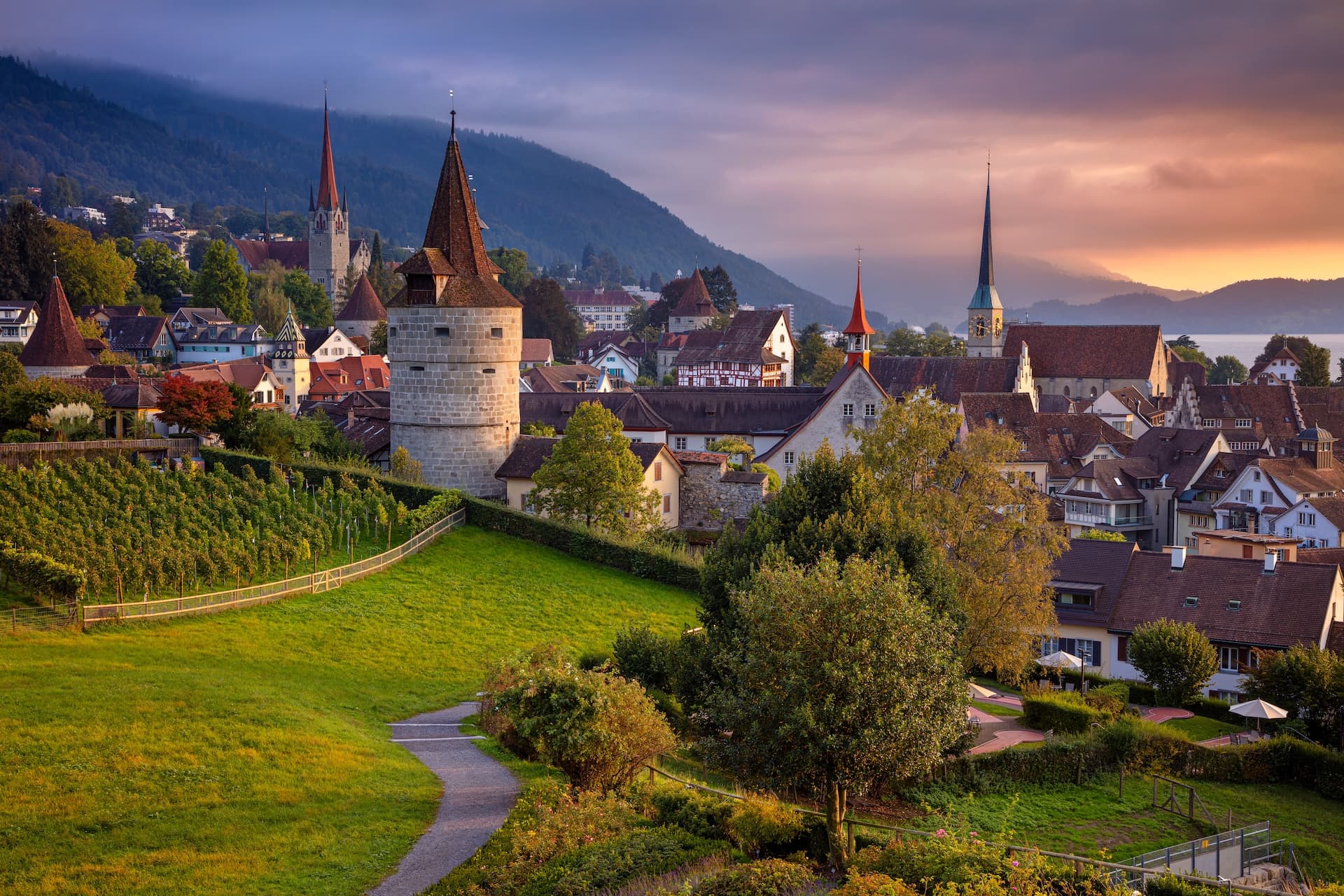 Zug, Switzerland cityscape with medieval tower, vineyards, and mountains at autumn sunset.