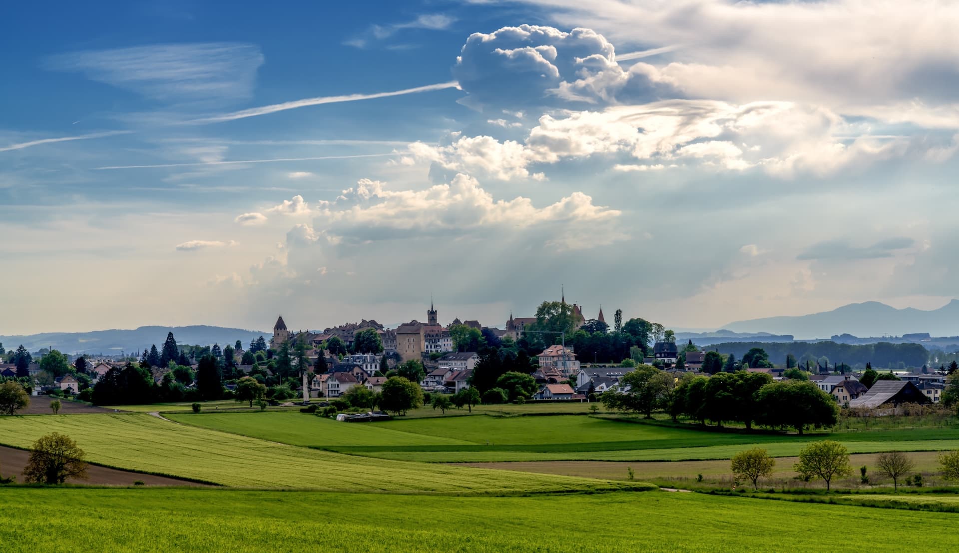 Historic town of Avenches above green fields under a dramatic, cloudy blue sky in western Switzerland.