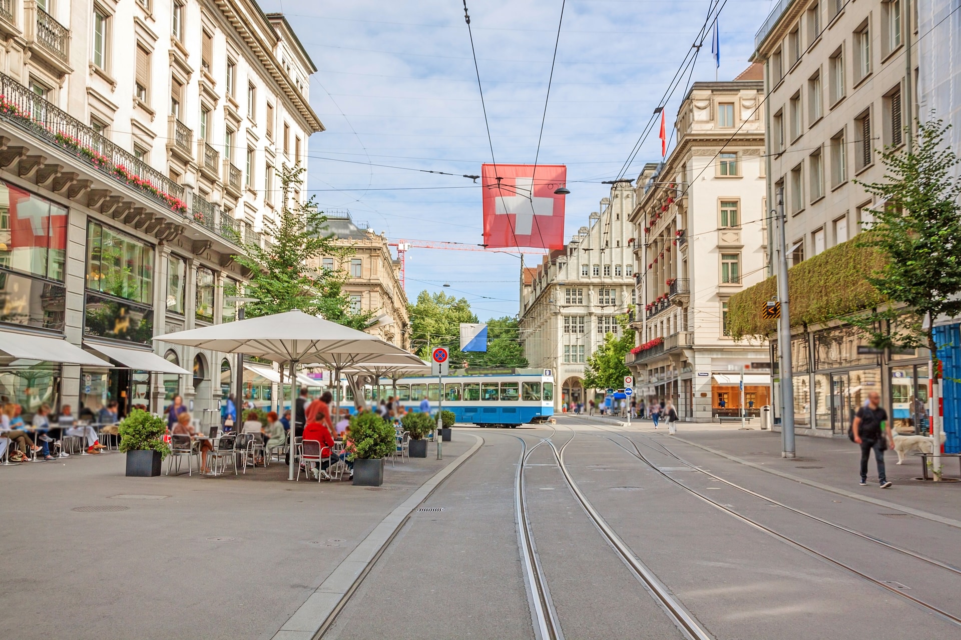 Tram on Bahnhofstrasse in Zurich with Swiss flag banner and outdoor cafe seating.