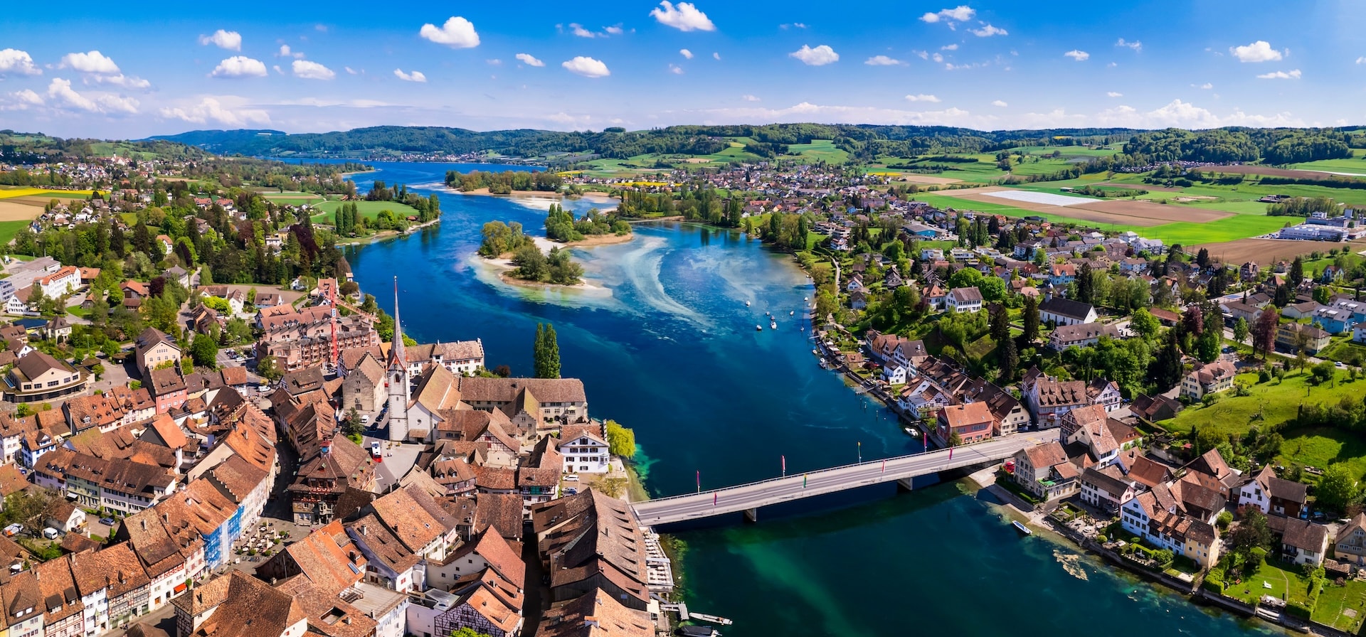Aerial view of Stein am Rhein old town, river, bridge, and green rolling hills under blue sky.